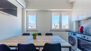 A modern kitchen with a built-in washing machine, a dining area with a table and chairs, and large windows offering a city view.