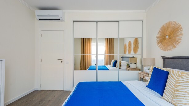 A hotel room with a large bed, a wardrobe with a mirror, a wall-mounted air conditioner, and a decorative wall piece.