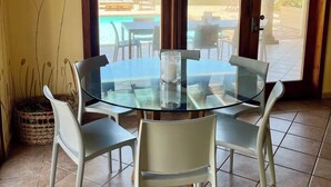 A dining area with a glass table and chairs, a hanging light fixture, and a view of the ocean through large sliding doors.