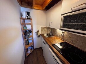 A compact kitchen with a built-in stove, a sink, and a wooden shelf unit with kitchen utensils.