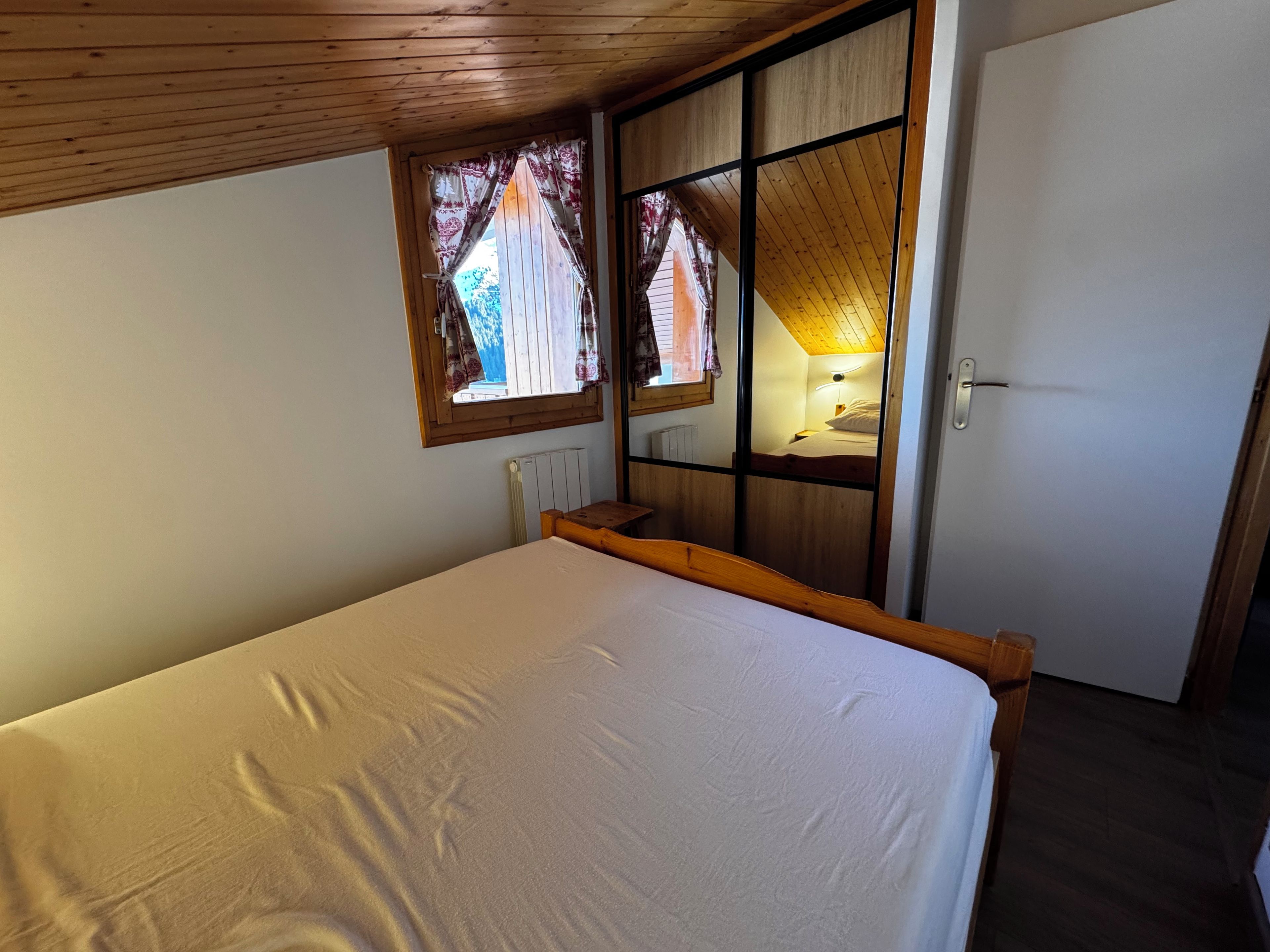 Une chambre avec un plafond en bois, un lit avec un matelas blanc, une fenêtre avec des rideaux et une porte.
