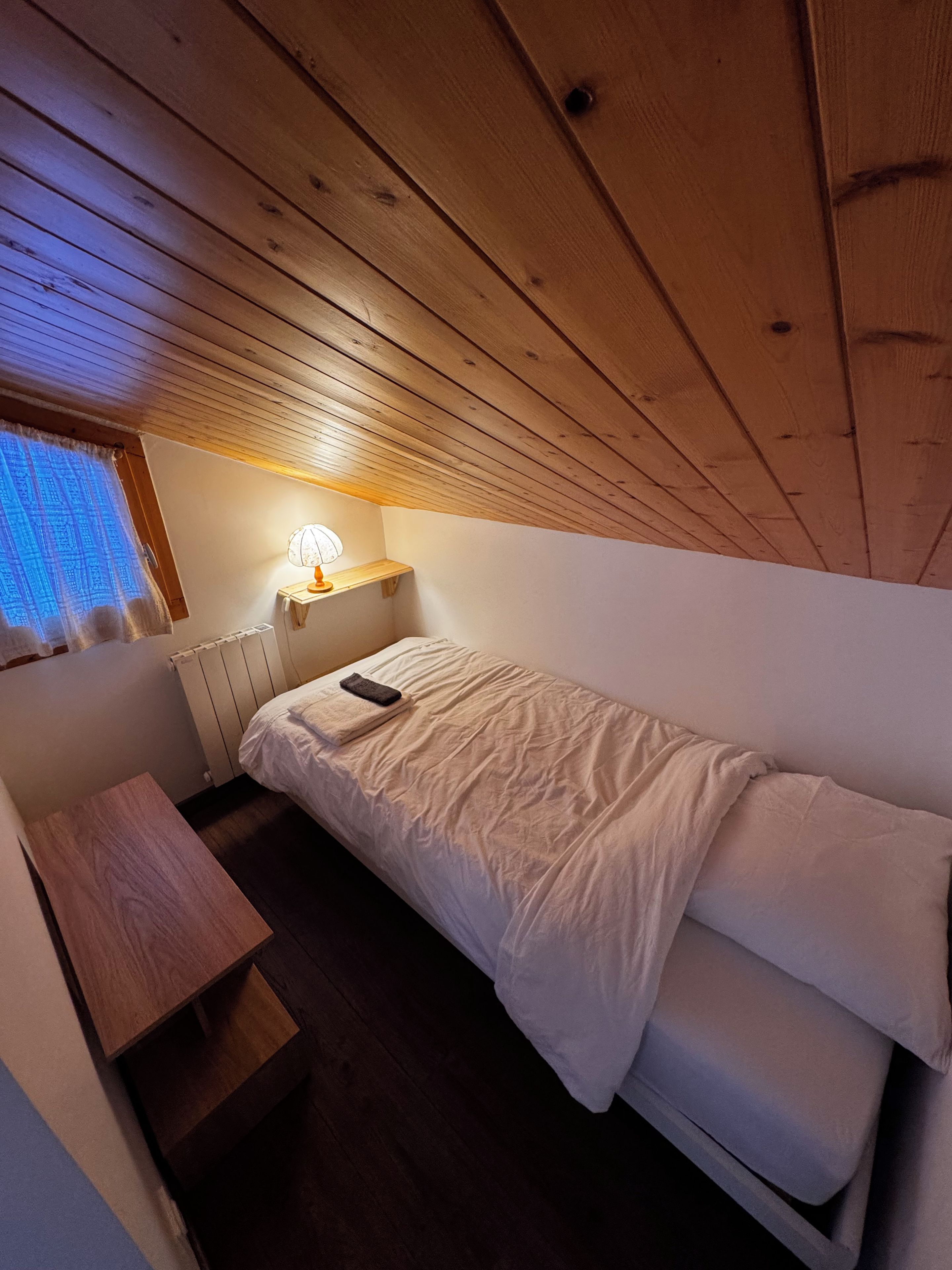 Ein kleiner, gemütlicher Raum mit einem Einzelbett, einem Nachttisch, einer Lampe und einem Fenster mit Vorhängen.