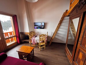 A room with a wooden floor, a television mounted on the wall, a wooden table, two chairs, a sofa, and a large mirror.