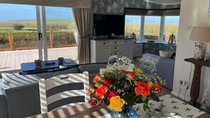 A room with a large window, a television, a dining table with chairs, and a floral arrangement. - Superior Stays at Sunset Lodge (Ilfracombe)