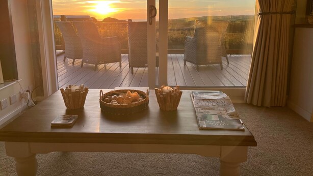A room with a wooden table, wicker chairs, and a view of the sunset through large windows. - Superior Stays at Sunset Lodge (Ilfracombe)