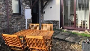 A patio with a wooden table and chairs, a stone wall, and a sliding glass door. - Braunton Breaks - Ambrose Cottage (Braunton)