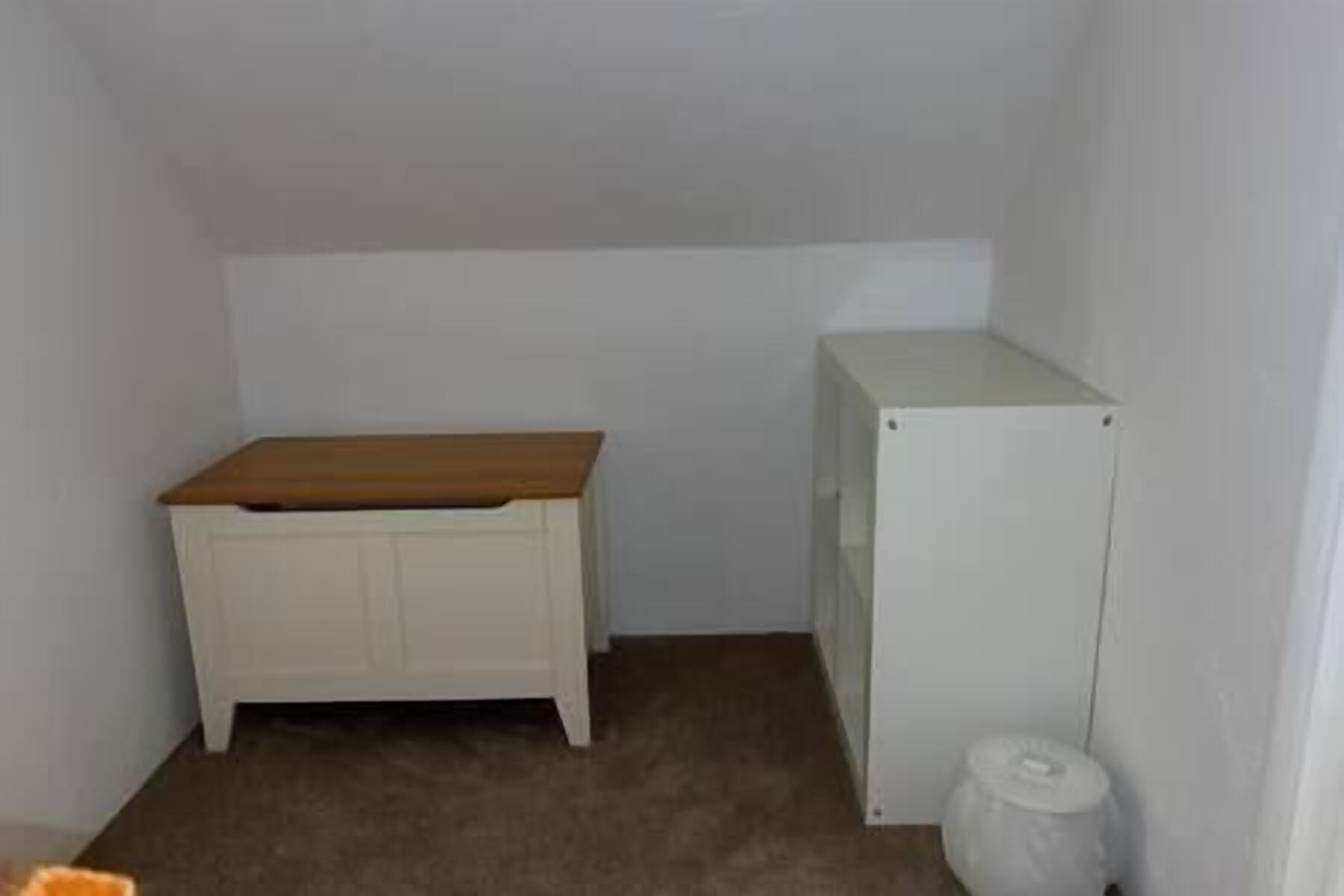 Um quarto pequeno, com pouca luz, com um banco de madeira e um armário branco.