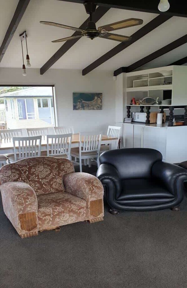 Un salon avec un canapé à motifs, un fauteuil en cuir noir, un ventilateur de plafond et un coin repas comprenant des chaises blanches et une table.