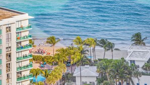 Un hotel frente a la playa con varios balcones, piscina y zona de playa con palmeras.