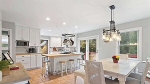 Une cuisine moderne avec des meubles blancs, un îlot central doté de tabourets, un porte-ustensiles suspendu et un coin repas avec une grande table et des chaises.