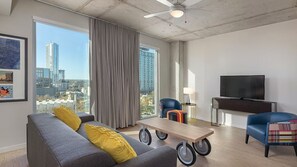Ein modernes Wohnzimmer mit grauem Sofa, gelben Kissen, einem Holz-Couchtisch auf Rollen, einem blauen Sessel, einem Fernseher und einem großen Fenster mit Blick auf die Stadt.