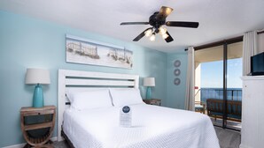 A bedroom with a bed, two bedside tables, a ceiling fan, a painting, and a view of the outdoors.