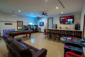 Une pièce équipée d’une table de billard, d’un canapé en cuir, de jeux d’arcade, d’un téléviseur diffusant des services de streaming et d’un ventilateur de plafond.