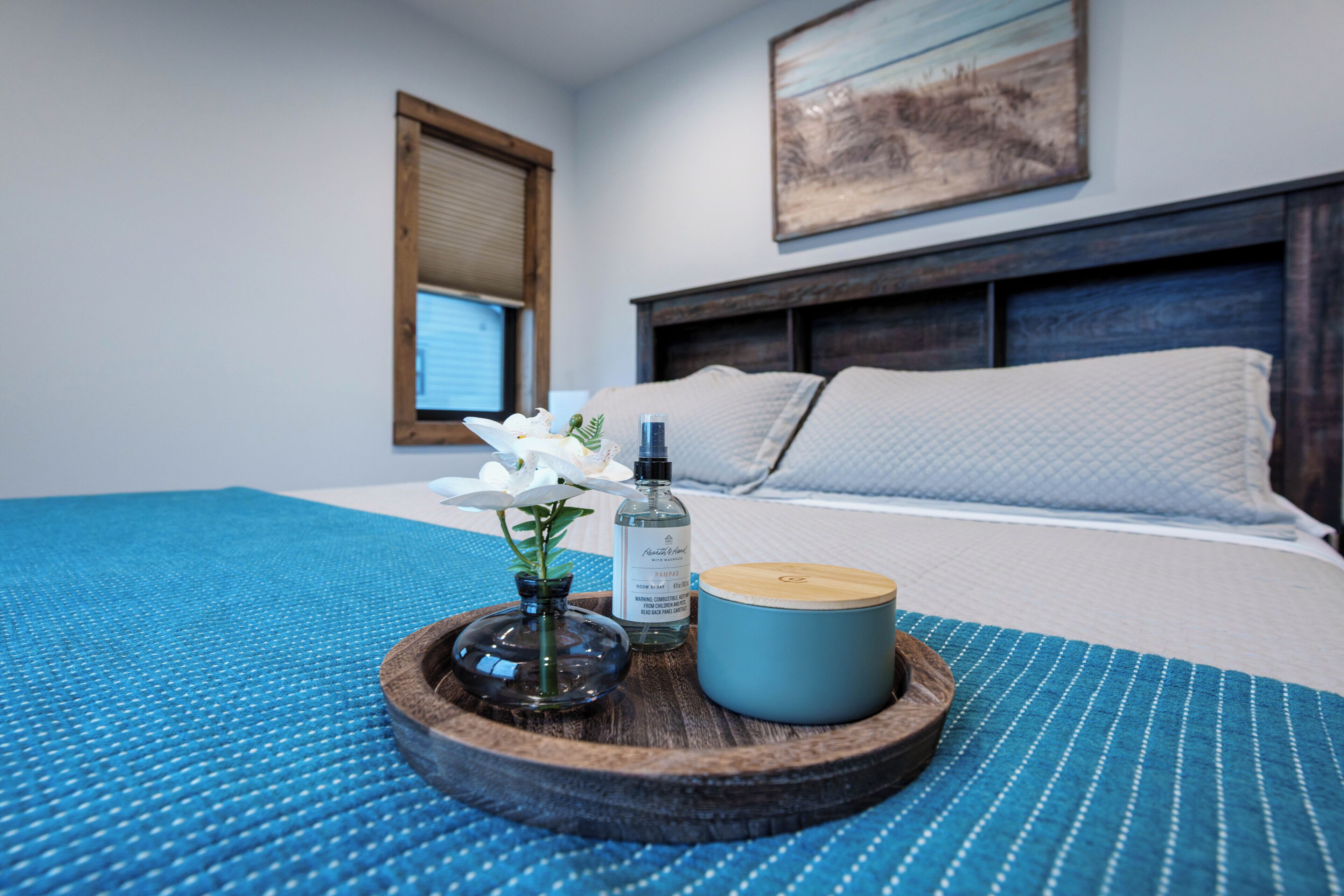 Ein ordentlich bezogenes Bett mit einer blauen, strukturierten Tagesdecke, ein Holztablett mit einer Kerze, einer Flasche und einer Blumenvase.