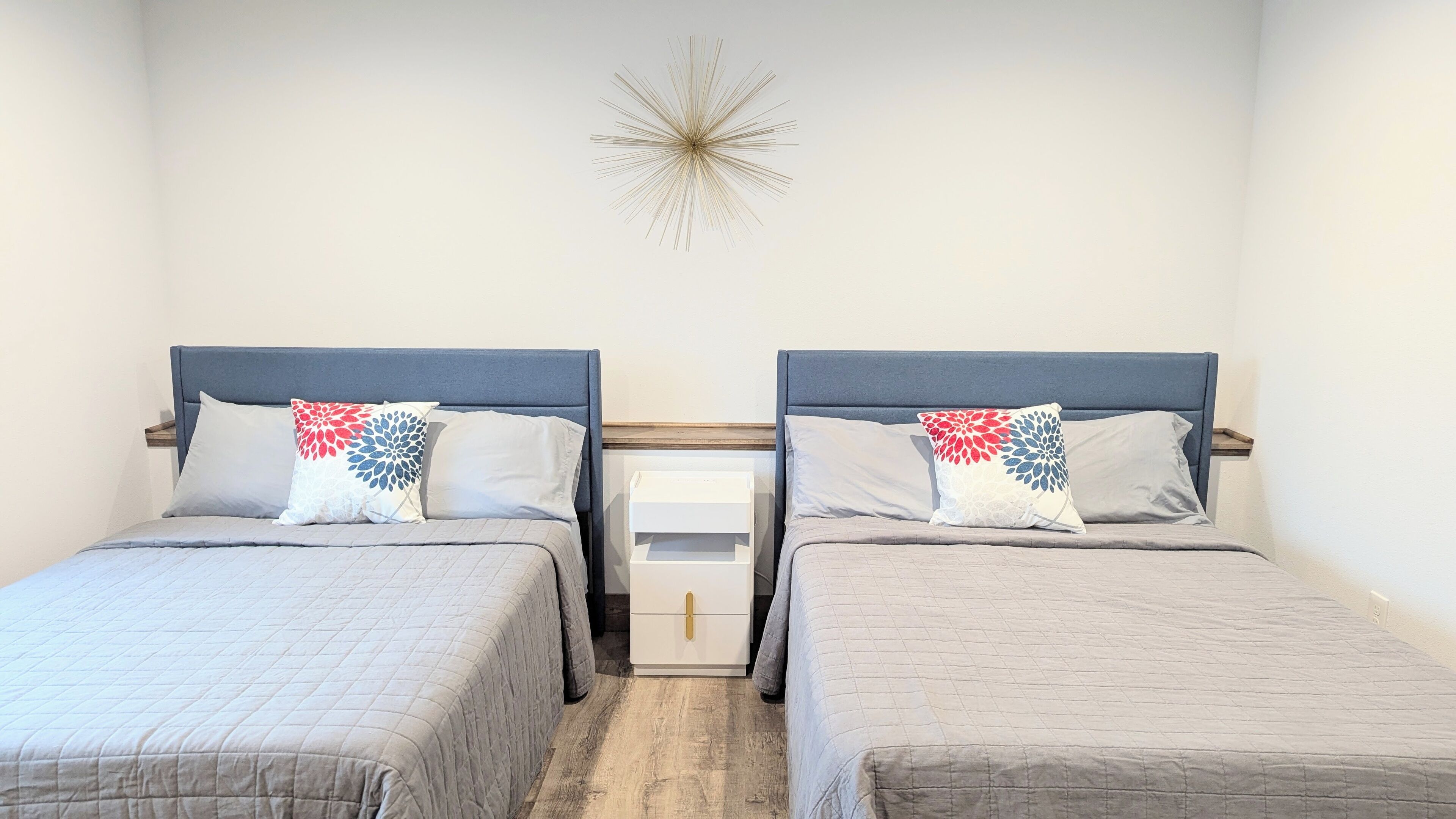 Ein Hotelzimmer mit zwei Betten, jeweils mit grauem Kopfteil und Nachttisch in Weiß. An der Wand über den Betten ist ein dekorativer Stern angebracht.