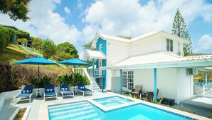 Ein Swimmingpool mit Liegestühlen und Sonnenschirmen, eine überdachte Terrasse und ein zweistöckiges Gebäude mit weißer Fassade.