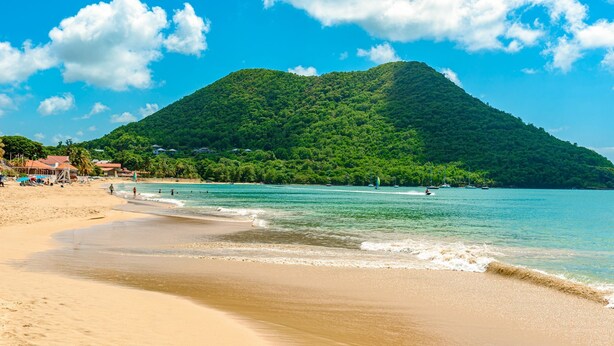 Ein Strand mit klarem türkisblauem Wasser, einem sandigen Ufer und einem üppigen, grünen Hügel im Hintergrund.