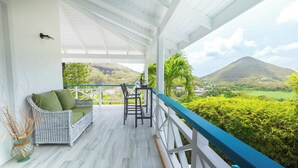 Eine überdachte Veranda mit Rattanmöbeln, einem Esstisch und Blick auf eine grüne Landschaft mit einem Berg.