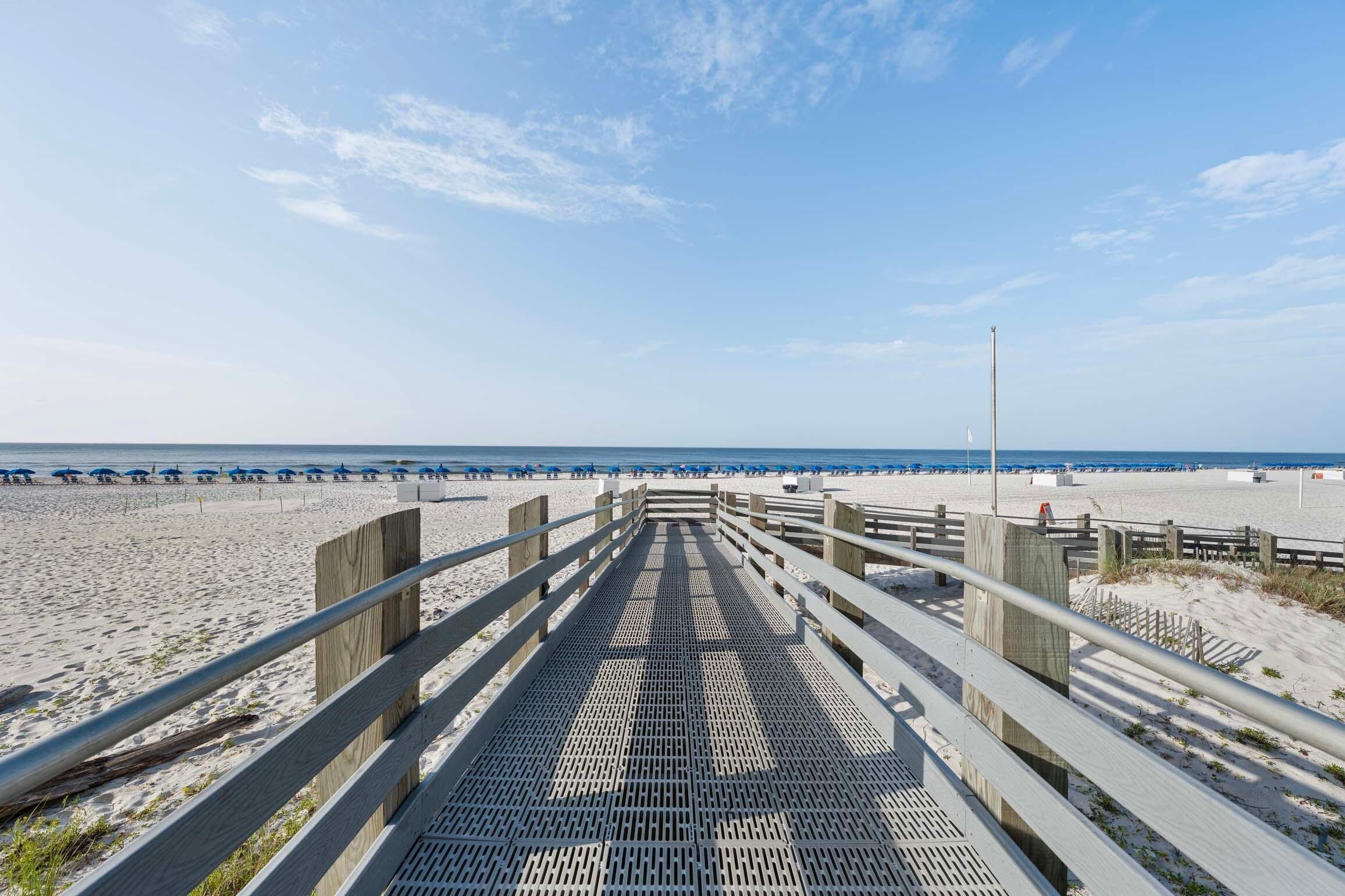 A wooden boardwalk leading to a beach with umbrellas and beachgoers.