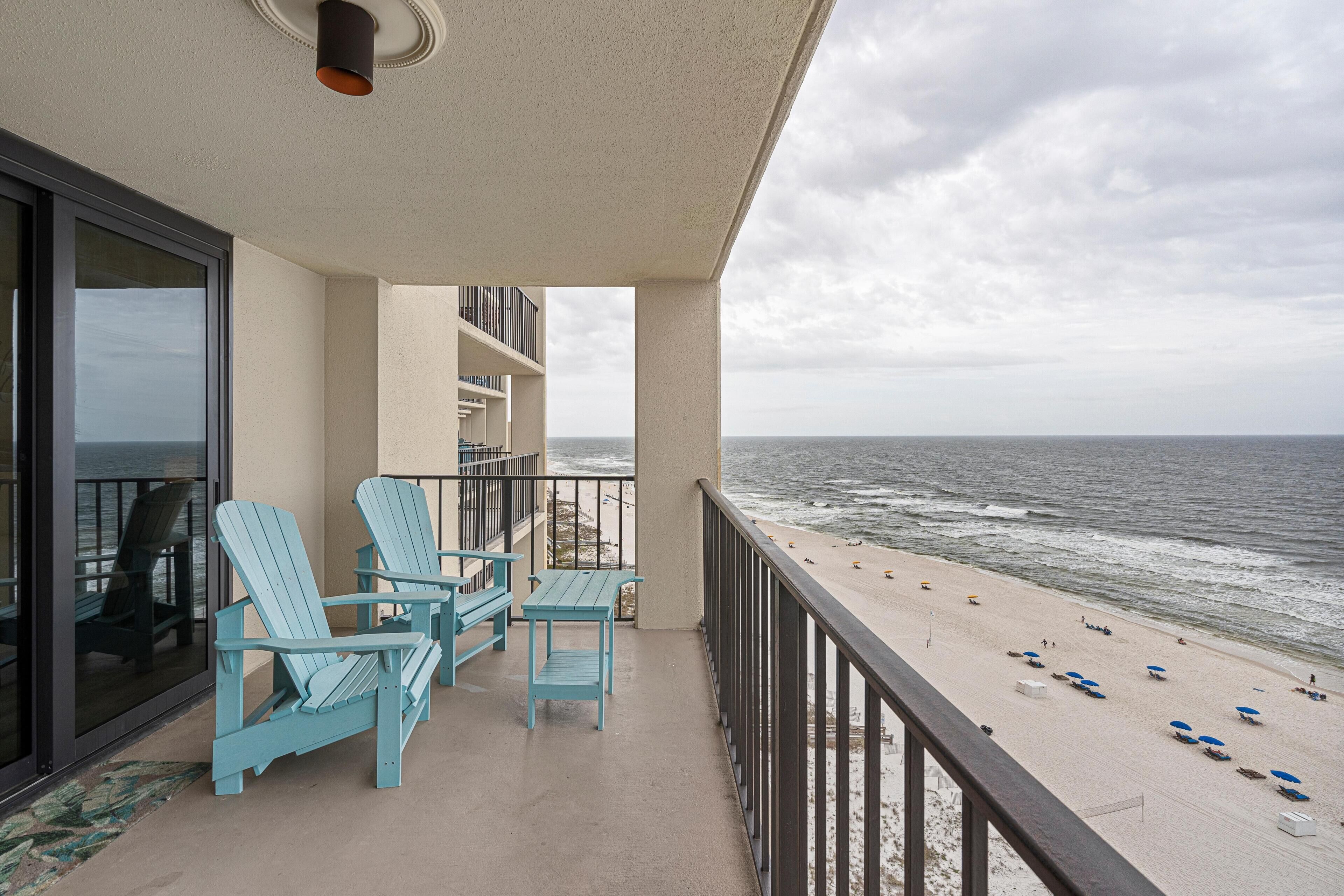 Ein Balkon mit blauen Adirondack-Stühlen und einem Tisch, mit Blick auf einen Strand mit Sonnenschirmen und Menschen.