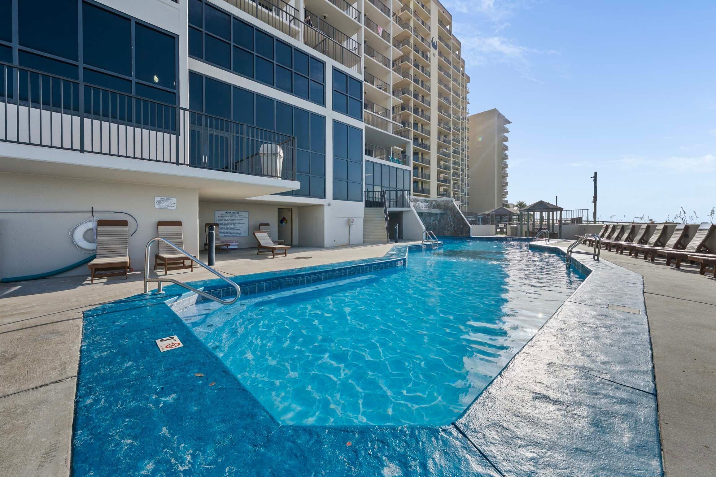 A swimming pool with a poolside area, lounge chairs, and a multi-story building in the background.