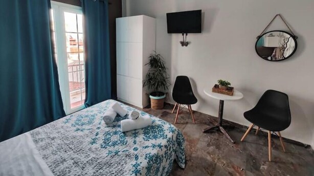 A hotel room with a bed, a TV, a small table with chairs, a potted plant, and a view through a window with curtains.