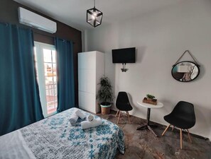 A hotel room with a bed, a TV, a small table with chairs, a potted plant, and a view through a window with curtains. - Blue Dreams (Veria)