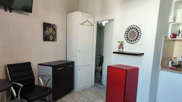 A compact room with a black office chair, a small black desk, a white wardrobe, a red refrigerator, and a wall-mounted mirror.