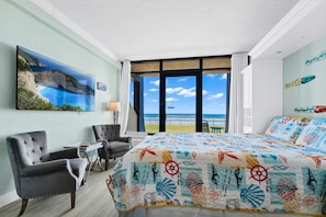 Une chambre d’hôtel avec un grand lit, deux fauteuils, une petite table, un tableau représentant un paysage côtier et une vue sur la mer par de grandes fenêtres.