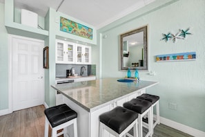 Une cuisine avec des meubles blancs, un îlot central en marbre doté de tabourets, un grand miroir et des œuvres d’art décoratives au mur.