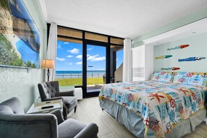 A bedroom with a bed, a chair, a table, a large window with a view, and a wall with decorative elements. - Stunning First Floor Direct Ocean Front Upgraded Studio SUMMER SALE POOL OPEN (Daytona Beach Shores)