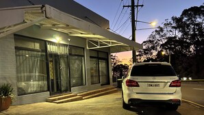 Un coche blanco estacionado frente a un edificio con ventanas grandes y un acceso cubierto.
