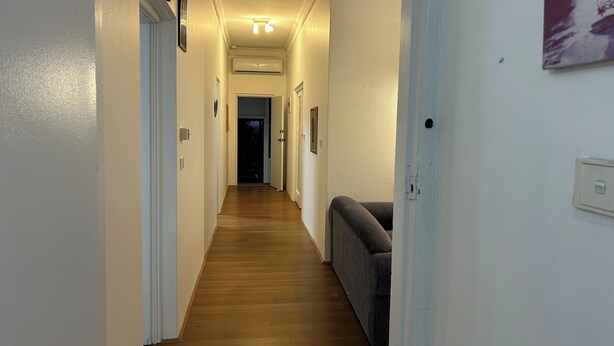 Un pasillo estrecho con suelo de madera, paredes blancas y un sofá gris.