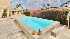 A swimming pool with a concrete border, surrounded by a garden with palm trees and pink flowers. - Beija Flor Entire home in Oranjestad, Aruba (Opal)
