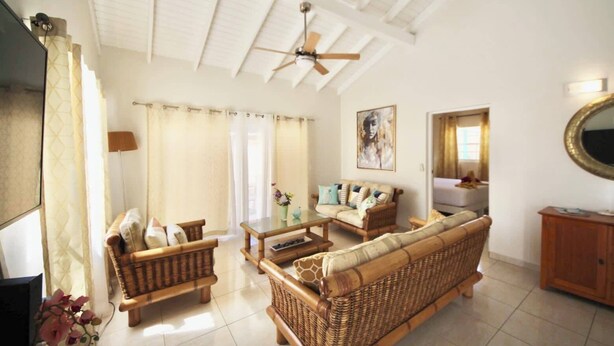 A spacious living room with a high ceiling, a ceiling fan, and a large window with curtains. There is a sofa, a coffee table, and a television. - Beija Flor Entire home in Oranjestad, Aruba (Opal)