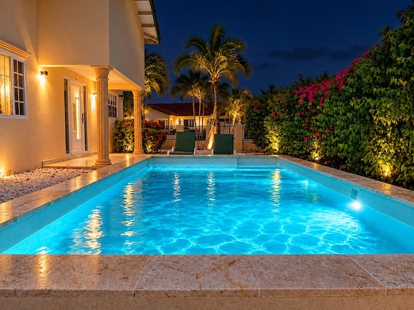 A swimming pool with a clear blue water surface, surrounded by a landscaped garden with palm trees and flowering bushes. - Beija Flor Entire home in Oranjestad, Aruba (Opal)