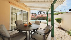 A covered outdoor dining area with a table and chairs, a glass door leading to a pool, and a view of a residential area with palm trees and a pink flower bush. - Beija Flor Entire home in Oranjestad, Aruba (Opal)