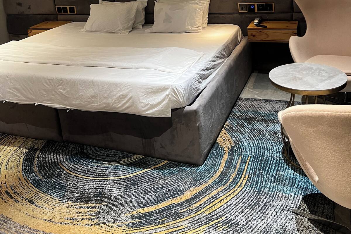 Una habitación de hotel moderna con una cama grande, un cabecero con listones de madera verticales, una obra de arte de mármol en la pared, una mesita redonda pequeña y una silla.