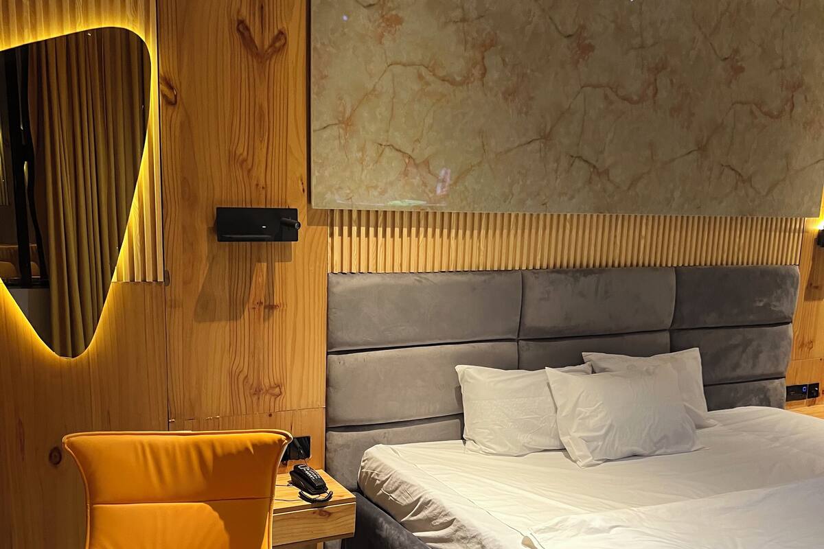 Una habitación de hotel moderna con una cama grande, cabecera de madera, una silla amarilla llamativa y una mesita de noche con un teléfono.