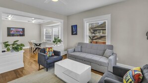 Ein geräumiges Wohnzimmer mit grauem Sofa, einem weißen Couchtisch und einem blauen Sessel. Es gibt einen Essbereich mit weißem Tisch und Stühlen sowie einen Deckenventilator.