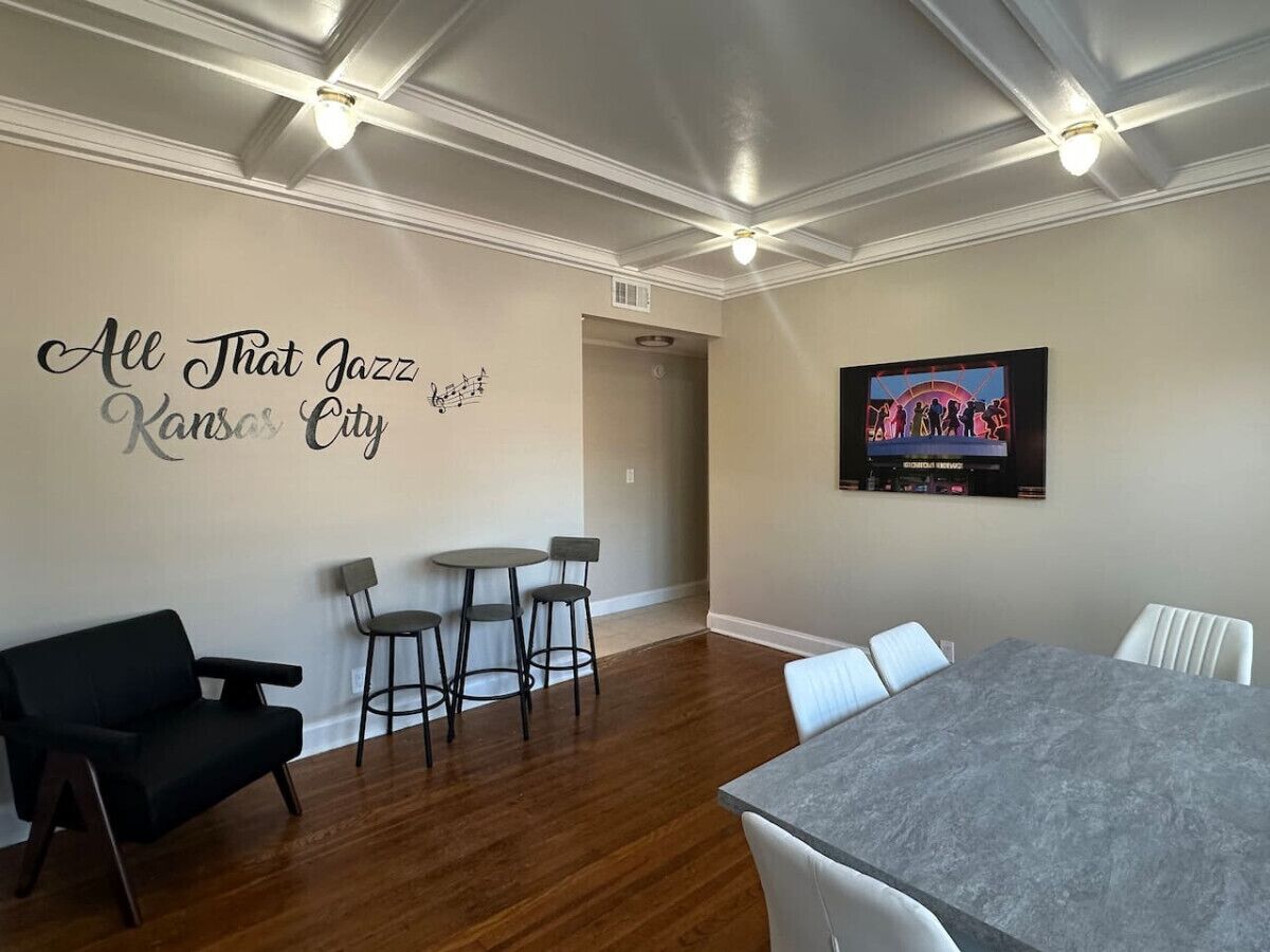 A modern living room with a black sofa, a dining table with chairs, a small kitchen area, and a wall with a decorative sign.