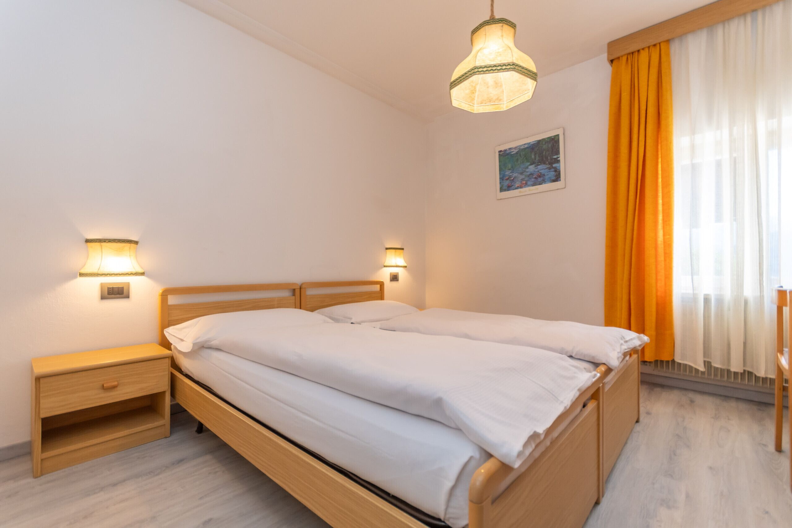 Un lit double avec deux tables de chevet, un luminaire suspendu, un tableau au mur et une fenêtre avec des rideaux.