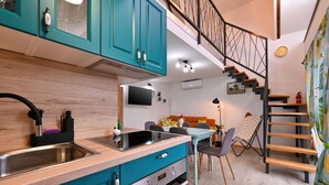 A modern kitchen with teal cabinets, a stainless steel sink, and a built-in oven. There is a dining area with a table and chairs, a TV mounted on the wall, and a staircase with a metal railing.