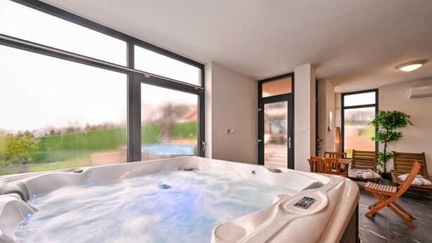 A modern spa room with a large hot tub, wooden seating, and large windows offering a view of the outdoors.