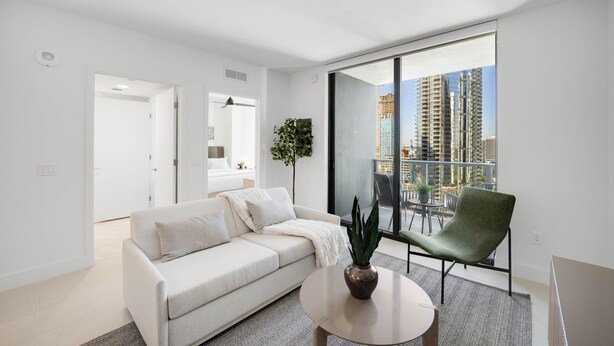 A modern living room with a sofa, a chair, a coffee table, and a large window offering a city view. - 601 RESIDENCES URBAN 1BR UNIT #2613 (Miami)