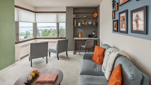 A modern living room with a grey sofa, orange cushions, a coffee table with a book, and a desk with a chair. There are framed pictures on the wall and a large window with blinds. - Artsy 1 Bedroom Condo with the Views to the Inlet! (Anchorage)