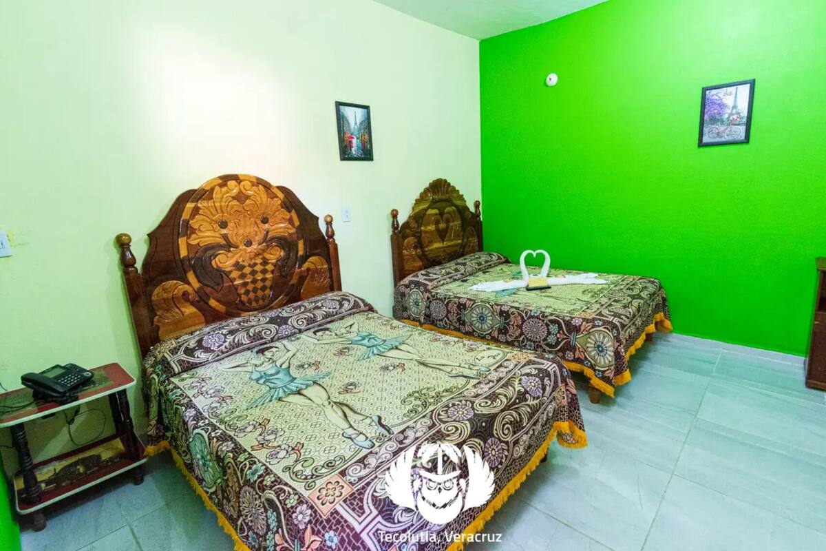 A room with two beds, each with a unique carved headboard, a small bedside table, and a green wall.