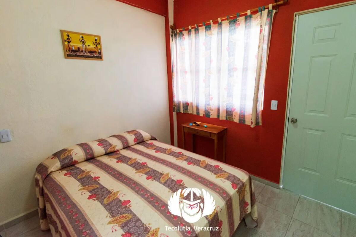 A bedroom with a bed, a small table, a window with curtains, and a door.