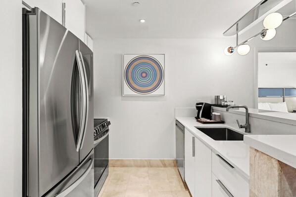 Una cocina moderna con un refrigerador de acero inoxidable, un lavamanos y una encimera. Se observa una obra de arte en la pared y una lámpara en el techo.