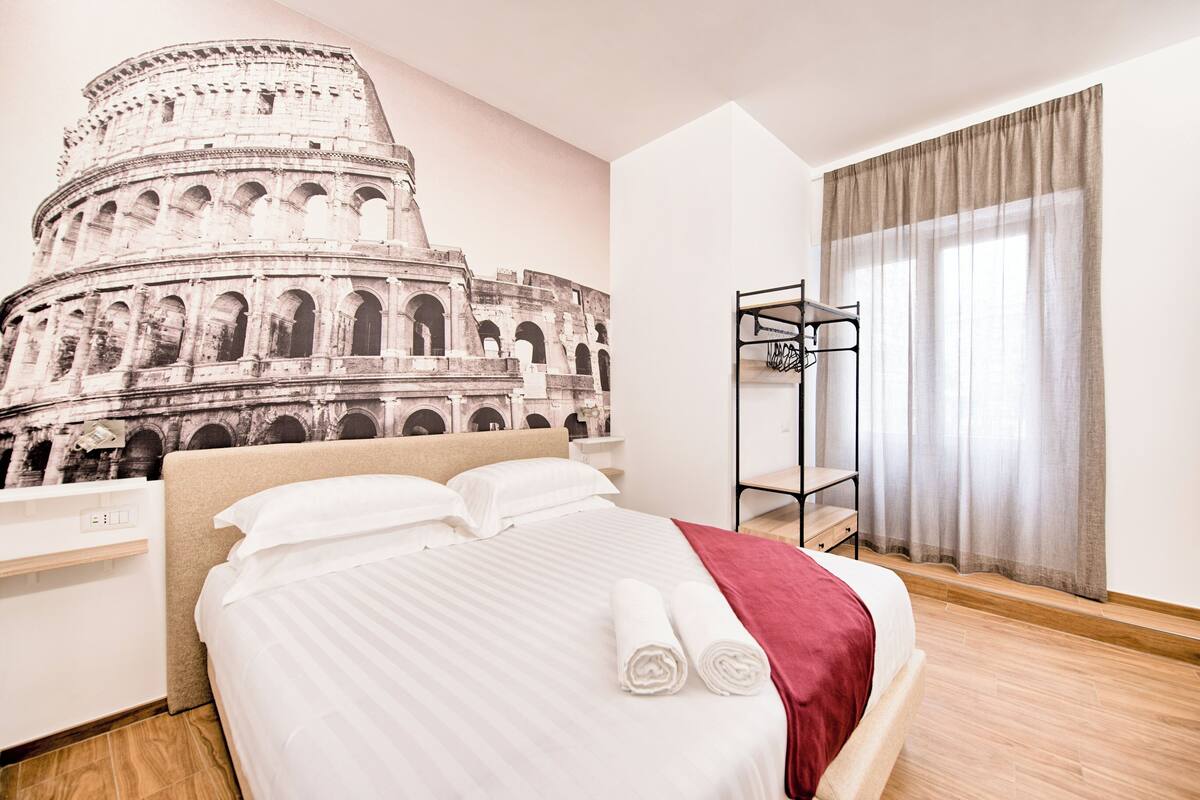 Ein Hotelzimmer mit Bett, Nachttisch, Wandregal und einem Fenster mit Vorhängen.